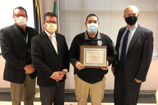 Four men stand in a row; three wear suits and one holds a framed certificate. All are wearing face masks.