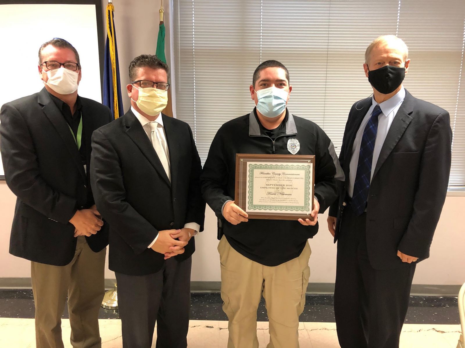 Four men stand in a row; three wear suits and one holds a framed certificate. All are wearing face masks.