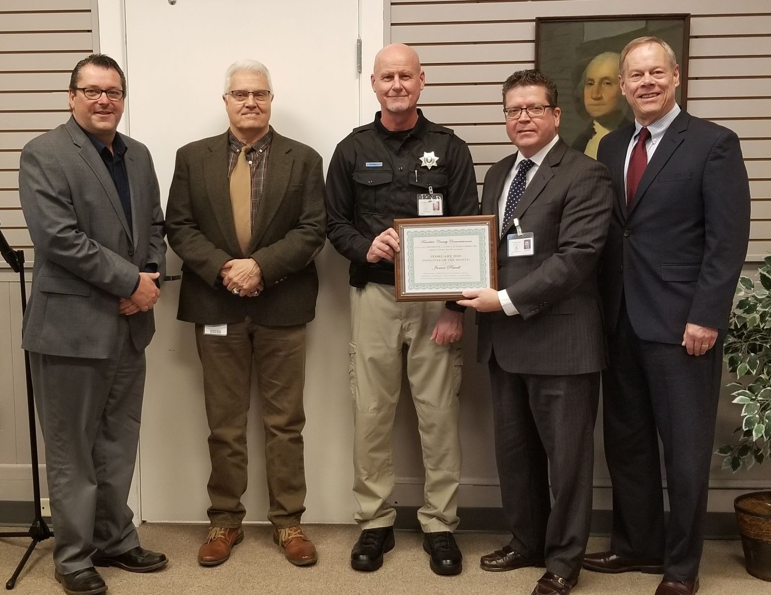 Five men in formal attire stand indoors; one holds a framed certificate.