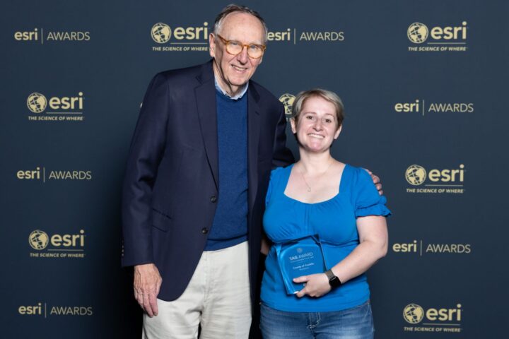 Two people stand together smiling at an Esri awards event. One is holding a plaque. The background displays the Esri logo and slogan 