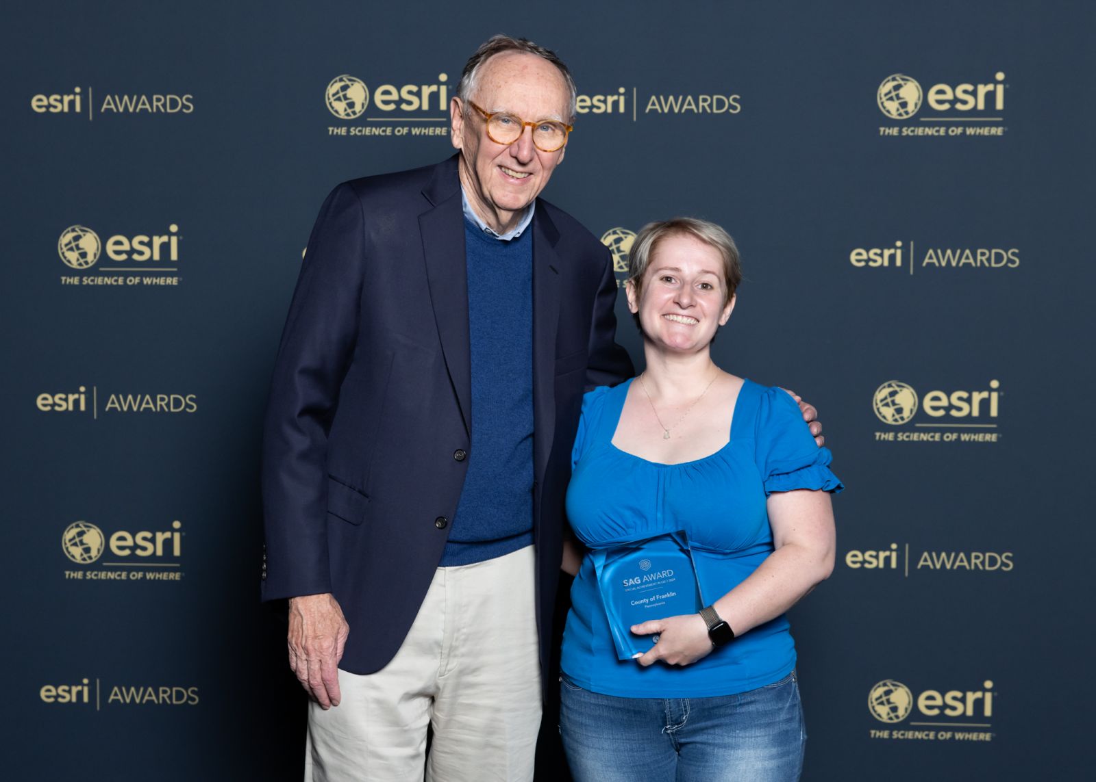 Two people stand together smiling at an Esri awards event. One is holding a plaque. The background displays the Esri logo and slogan 