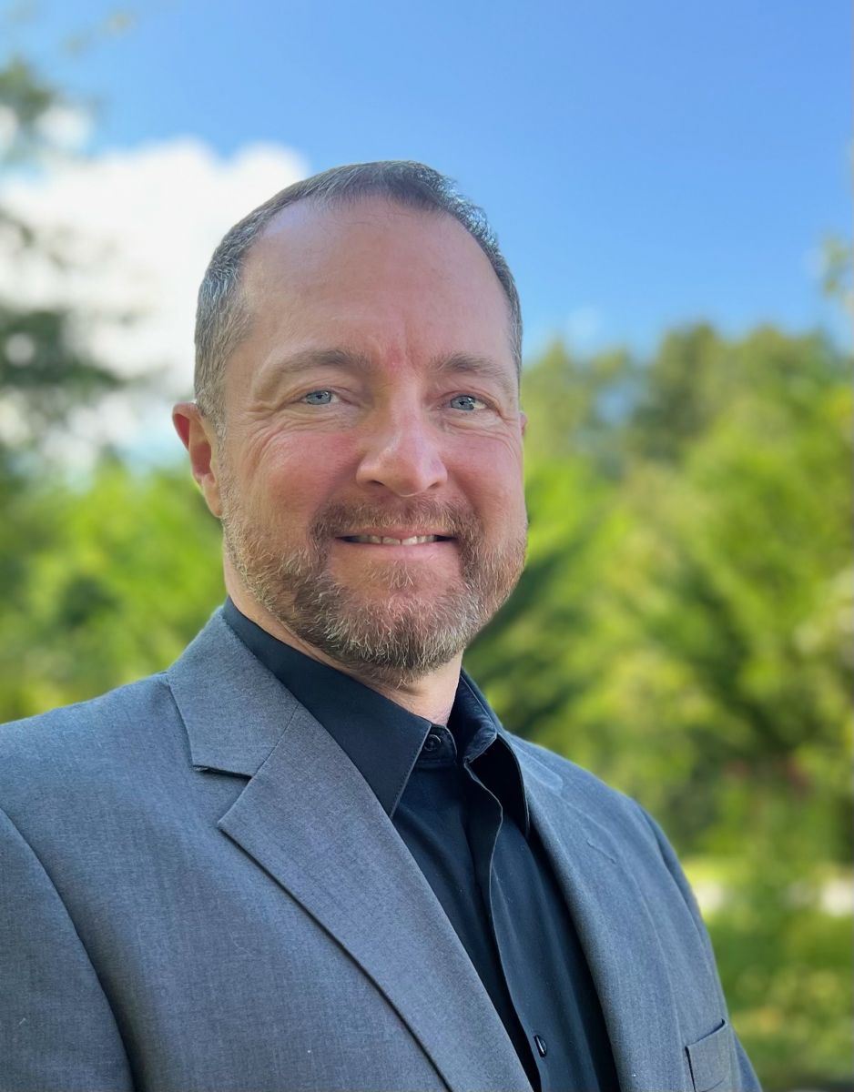 A man in a gray suit jacket and black shirt smiles at the camera with a blurred green and blue outdoor background.