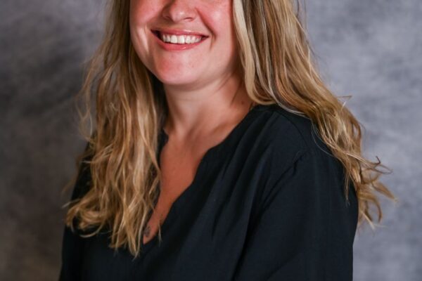 A woman with long blonde hair smiles while wearing a black shirt. The background is textured gray.