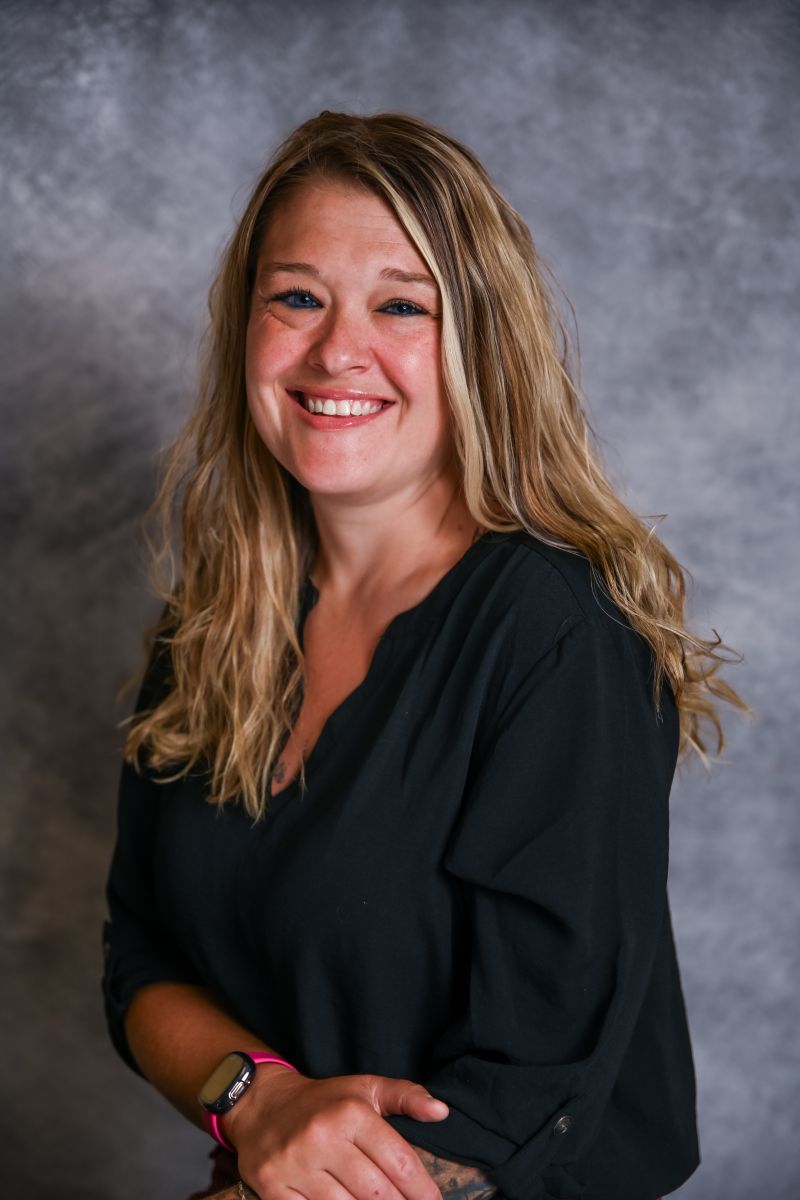 A woman with long blonde hair smiles while wearing a black shirt. The background is textured gray.