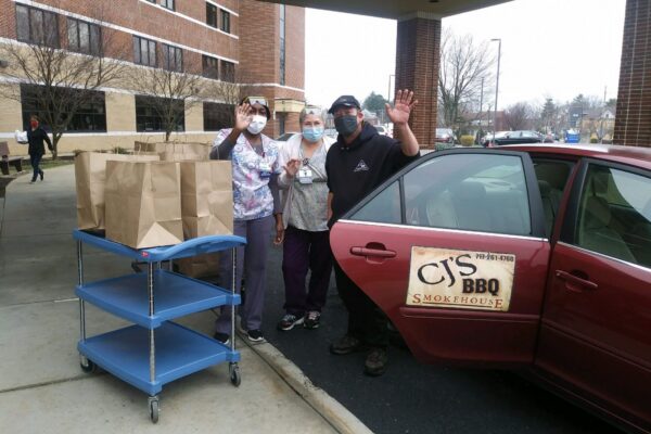 Three people wearing masks wave by a red car with 