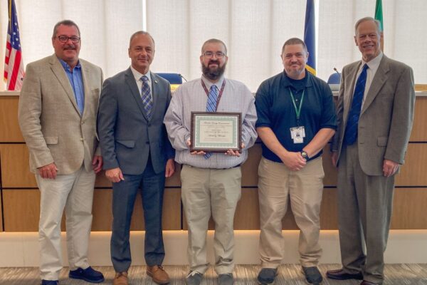 Five men standing together in a conference room; the person in the center holds a framed certificate.