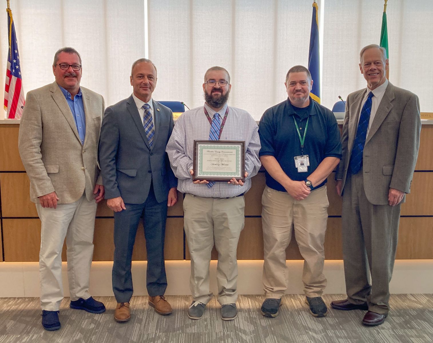 Five men standing together in a conference room; the person in the center holds a framed certificate.