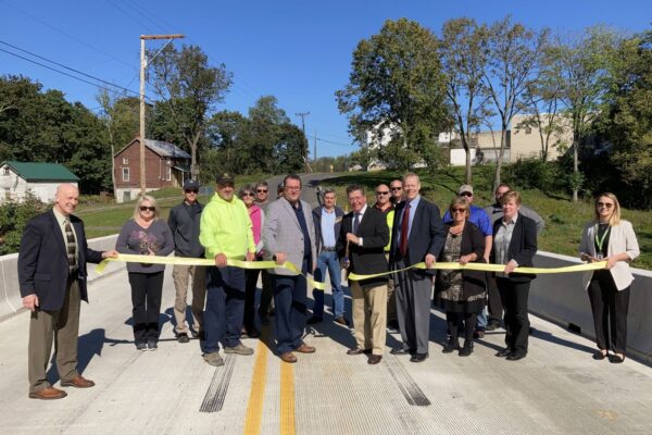 A group of people participate in a ribbon-cutting ceremony on a new bridge. Trees and buildings are in the background.