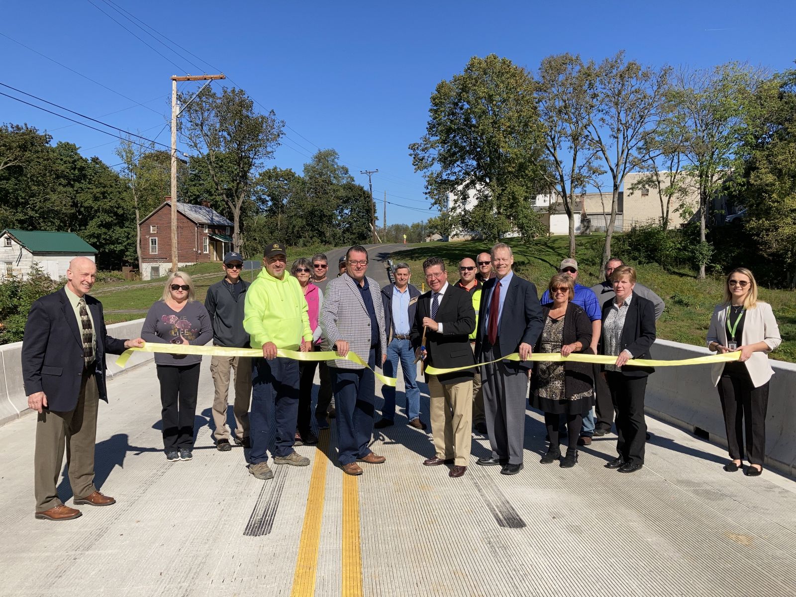 A group of people participate in a ribbon-cutting ceremony on a new bridge. Trees and buildings are in the background.