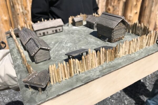 A person holds a small diorama of a historic wooden fort with several buildings inside a fenced perimeter.