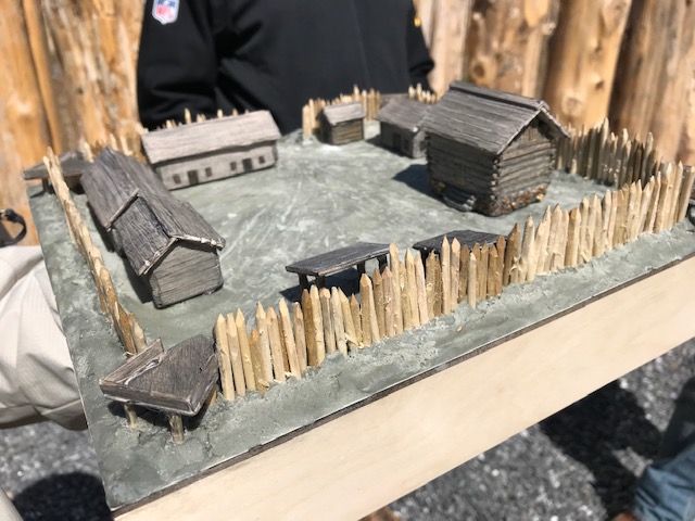 A person holds a small diorama of a historic wooden fort with several buildings inside a fenced perimeter.