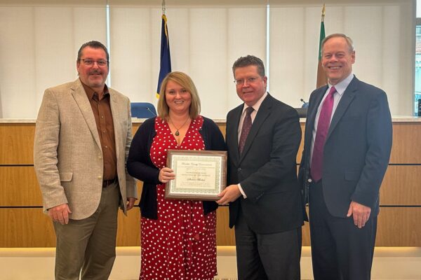 Four people stand together in a room, with one person holding a certificate. They are dressed in formal attire, and there are flags in the background.