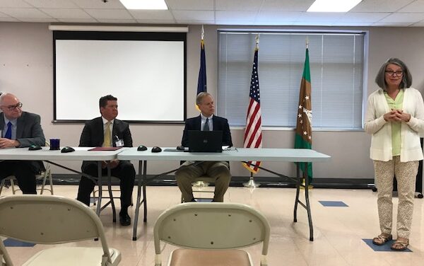 A woman stands speaking in a room with three men seated at a table. Behind them are flags and a projection screen.