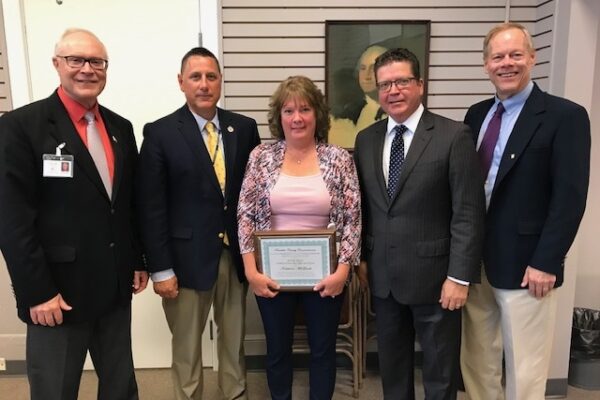 Five people in business attire stand together indoors. The person in the center holds a framed certificate.