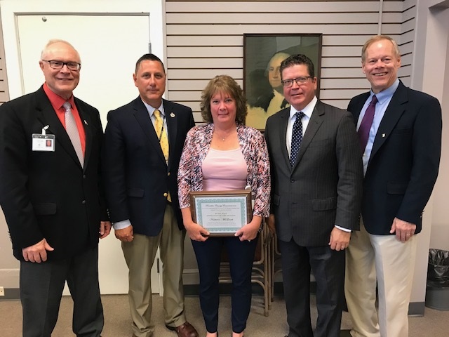 Five people in business attire stand together indoors. The person in the center holds a framed certificate.