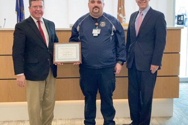 Three men stand in an office; the man in the middle holds a certificate.