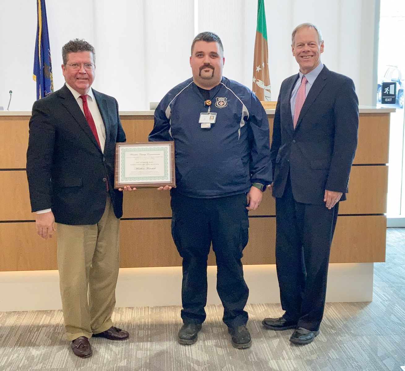 Three men stand in an office; the man in the middle holds a certificate.