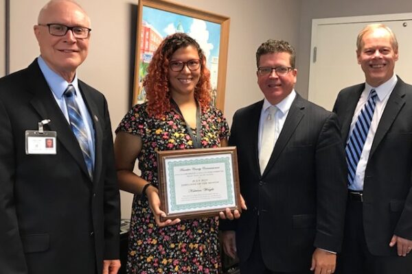 Four people are standing indoors. One person holds a framed certificate. All are dressed in business attire and smiling.