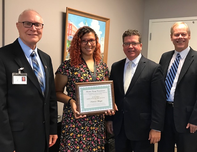 Four people are standing indoors. One person holds a framed certificate. All are dressed in business attire and smiling.