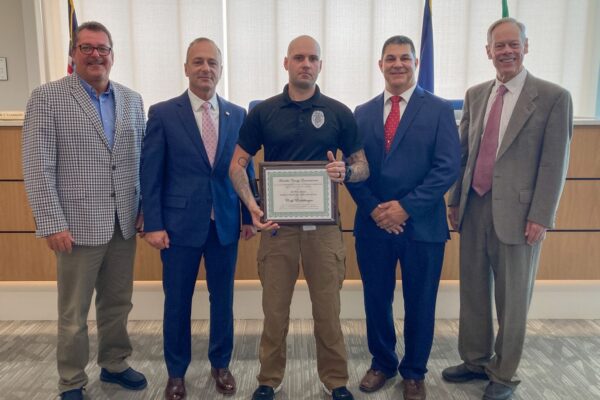 Five men stand indoors, with one in the center holding a framed certificate. They are dressed in business and business-casual attire. Flags are visible in the background.