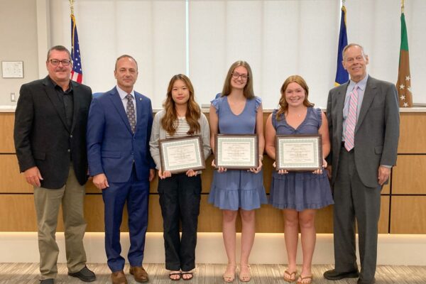 Six people stand indoors, with three young women in the center holding certificates. They are in professional attire, and flags are visible in the background.