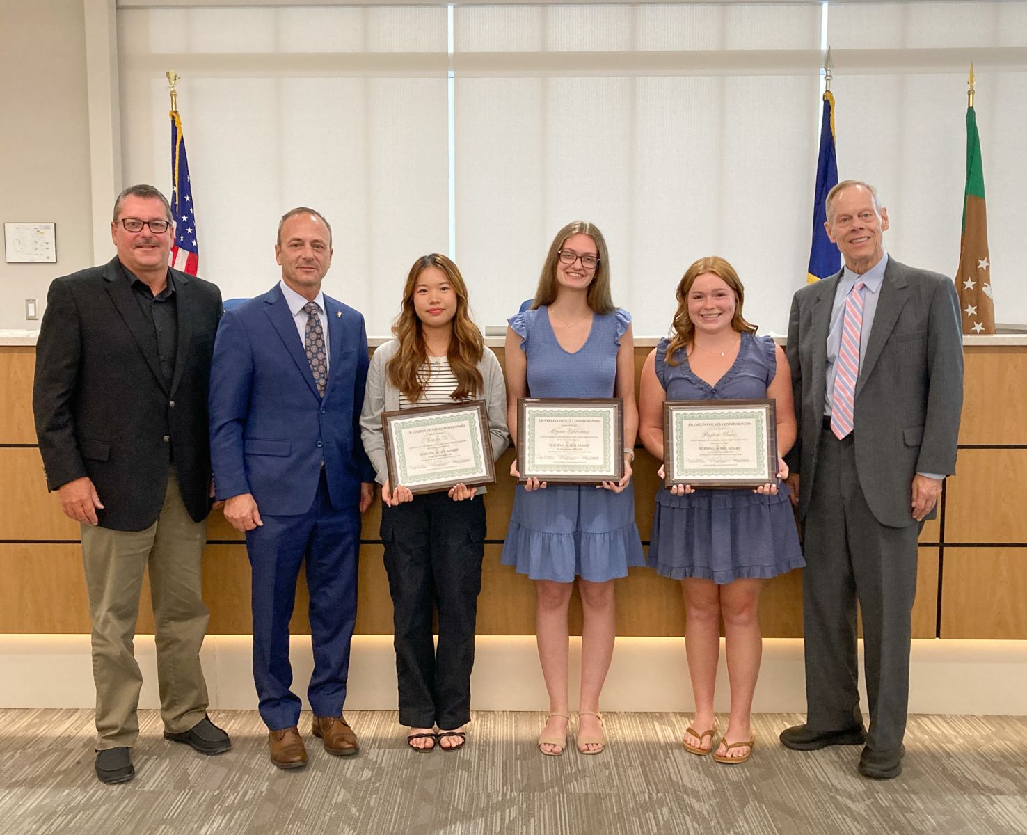 Six people stand indoors, with three young women in the center holding certificates. They are in professional attire, and flags are visible in the background.