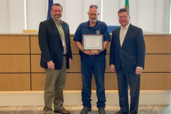 Three men stand together indoors; the middle man holds a framed certificate.