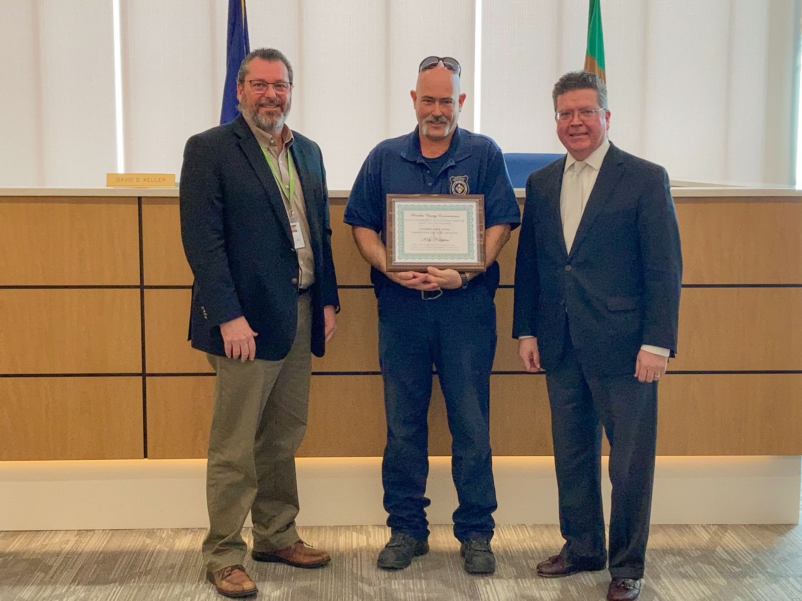 Three men stand together indoors; the middle man holds a framed certificate.