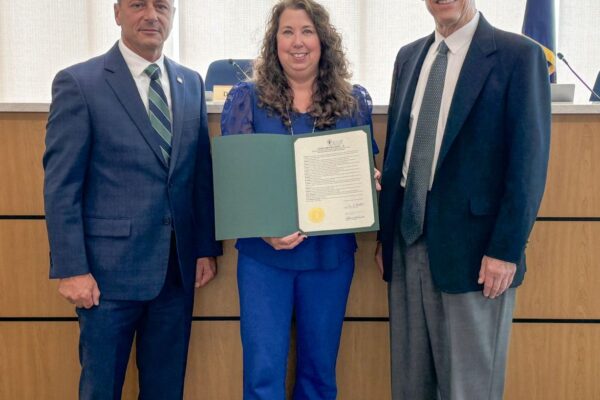 Three people stand in an office setting; the woman in the center holds a certificate. Two men in suits stand on either side.