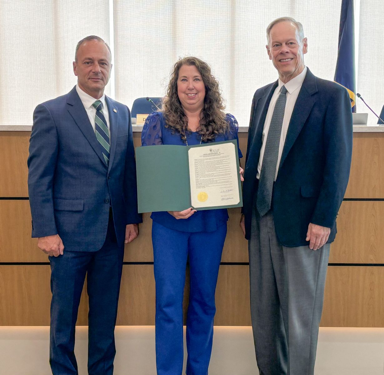 Three people stand in an office setting; the woman in the center holds a certificate. Two men in suits stand on either side.