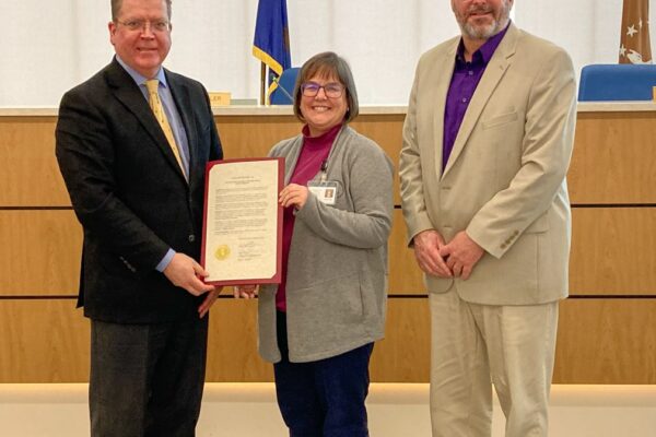 Three people stand indoors; one holds a certificate. Two men wear blazers, and the woman in the center holds a document. They are in a room with a flag and a seal in the background.