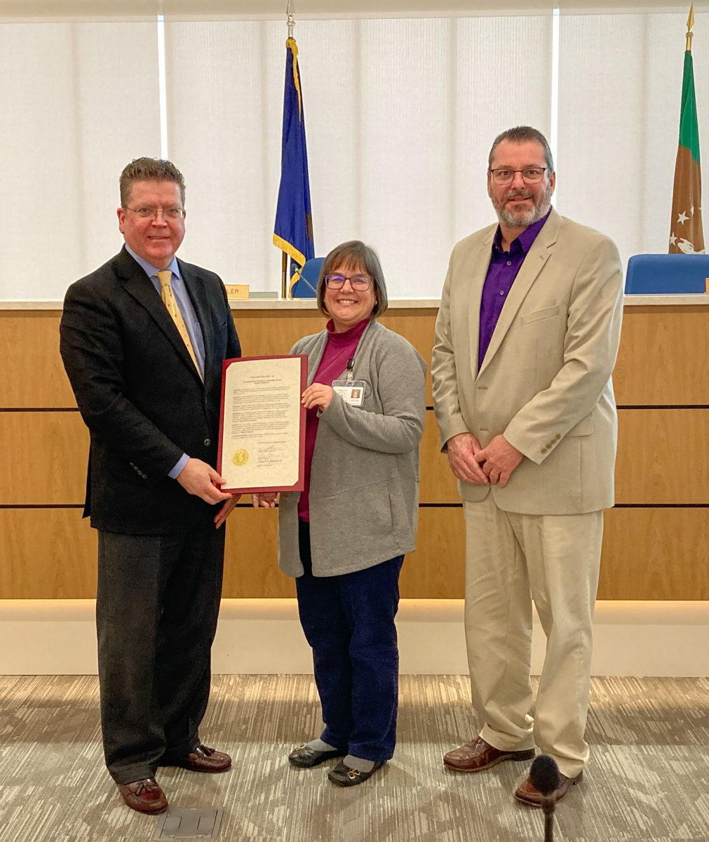 Three people stand indoors; one holds a certificate. Two men wear blazers, and the woman in the center holds a document. They are in a room with a flag and a seal in the background.