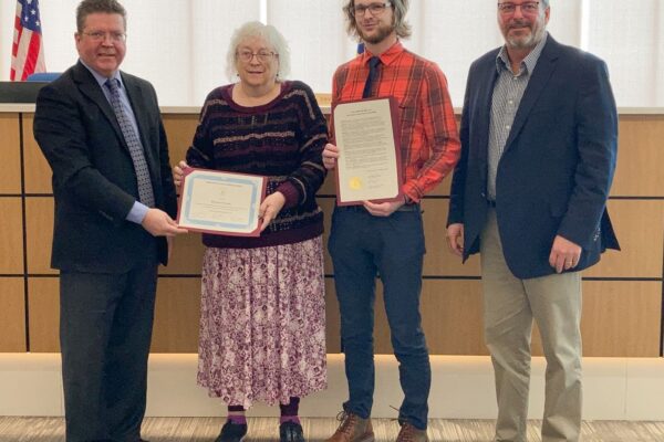 Four people standing in a room; two hold certificates.