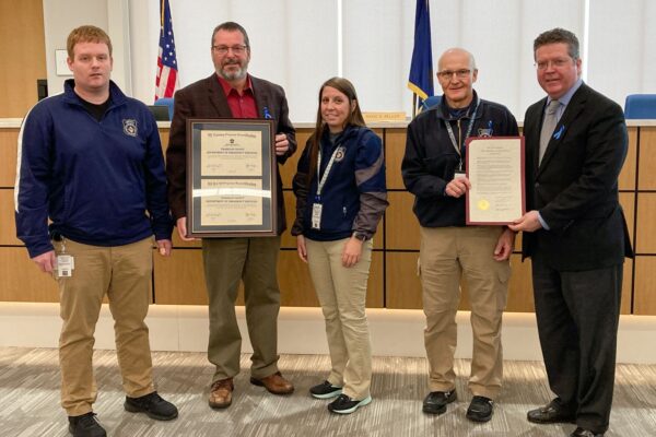 Five people stand indoors; two hold framed certificates. An American flag and government emblems are in the background.