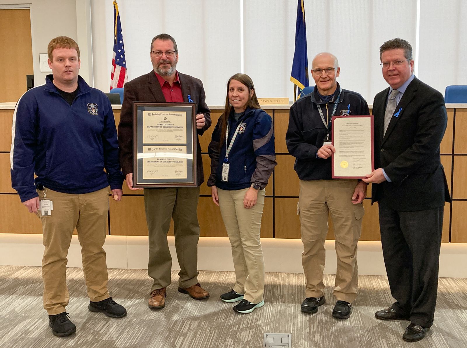 Five people stand indoors; two hold framed certificates. An American flag and government emblems are in the background.