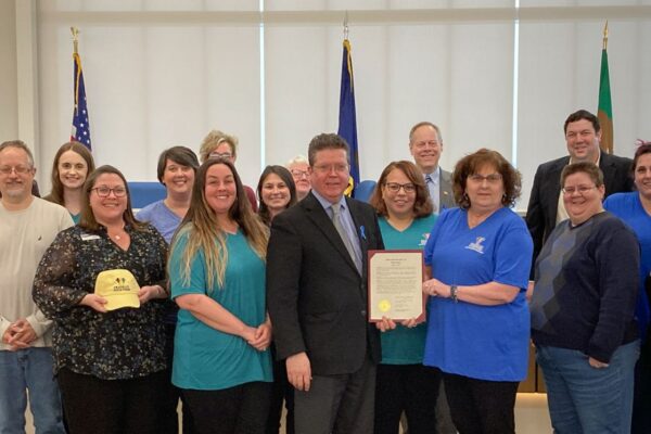 A group of people stands indoors, some smiling, with a man in the center holding a framed certificate. There are flags and a large window in the background.