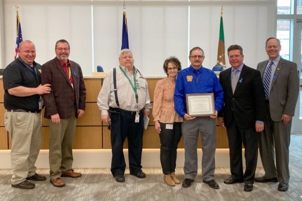 A group of people stand in an office room. One person holds a certificate. Flags are displayed in the background.