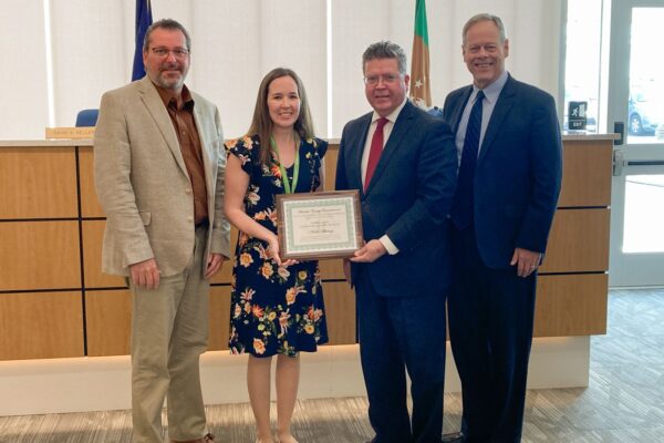 Four people stand indoors; one woman holds a framed certificate. They are dressed in business attire, and there is a wooden panel backdrop.