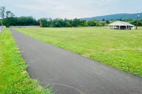 A paved path runs alongside a grassy field, with a pavilion in the distance under a cloudy sky. Trees and hills are visible in the background.