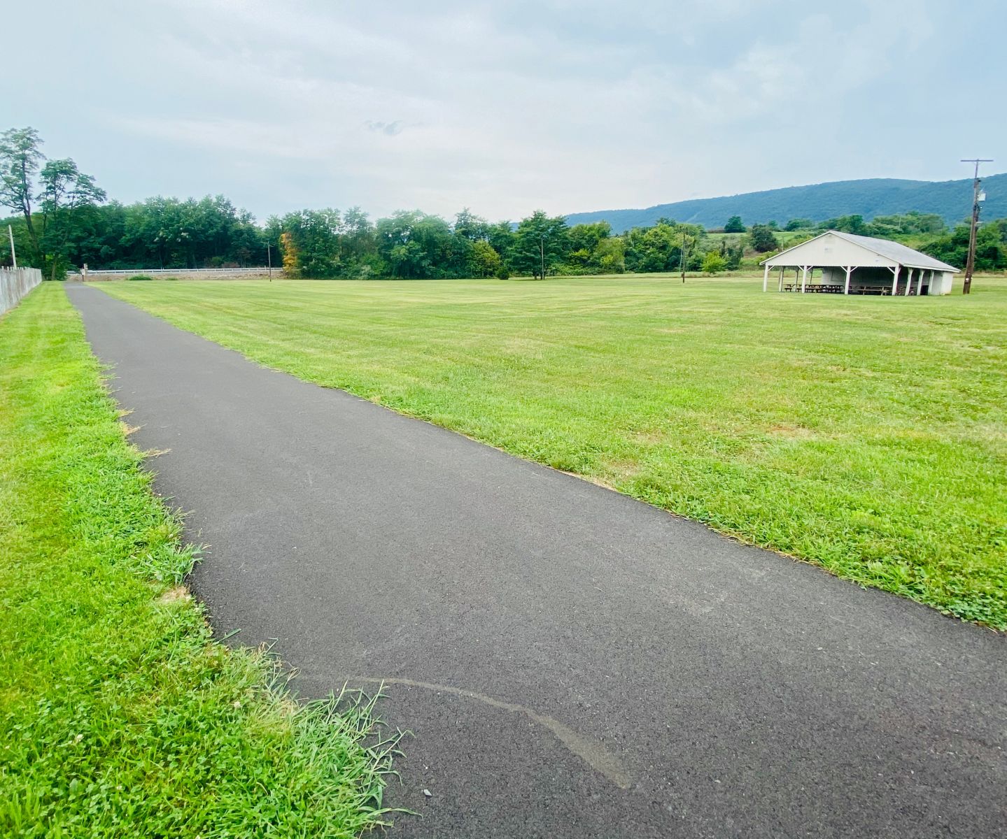 A paved path runs alongside a grassy field, with a pavilion in the distance under a cloudy sky. Trees and hills are visible in the background.