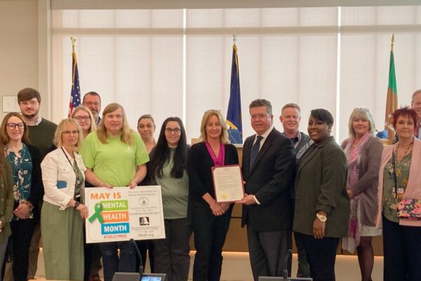 A group of people posed indoors, some holding a sign about May being Mental Health Awareness Month. There are flags in the background.