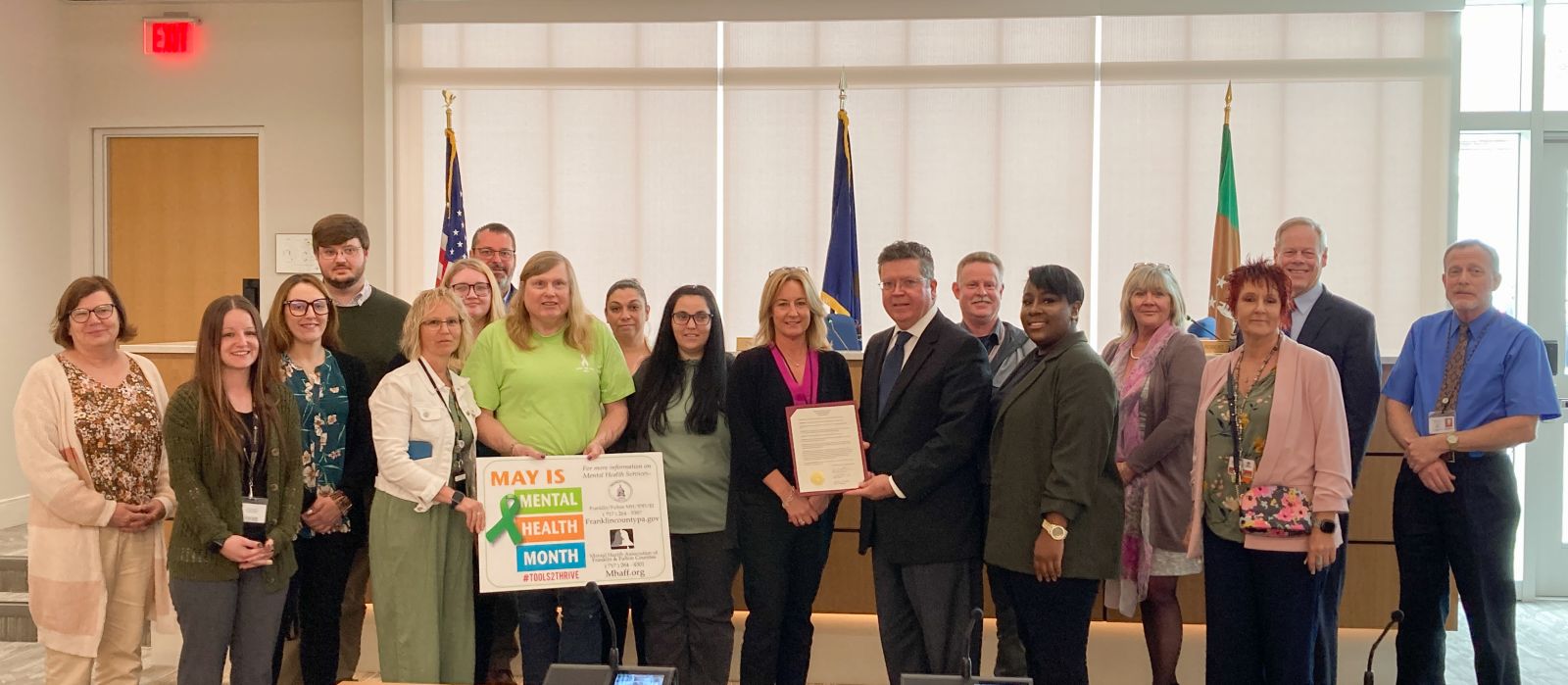 A group of people posed indoors, some holding a sign about May being Mental Health Awareness Month. There are flags in the background.