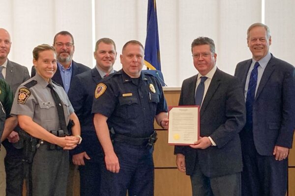 A group of people in formal attire, including law enforcement officers, pose indoors holding a framed document. Flags are visible in the background.