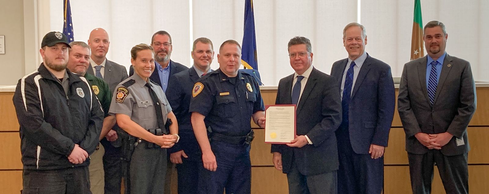 A group of people in formal attire, including law enforcement officers, pose indoors holding a framed document. Flags are visible in the background.