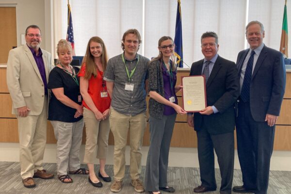 A group of seven people stand indoors, with one person holding a framed certificate. They are positioned in front of flags and a wooden panel backdrop.