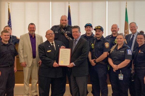 A group of people, including firefighters and officials, pose indoors holding a framed document. American flags are visible in the background.