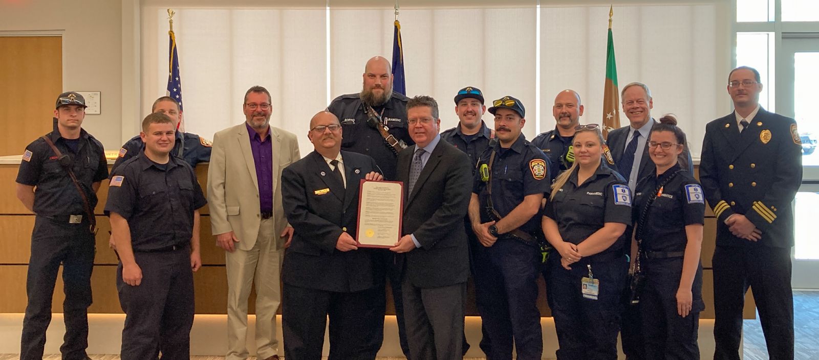 A group of people, including firefighters and officials, pose indoors holding a framed document. American flags are visible in the background.