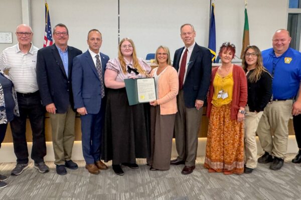 A group of eleven people standing indoors, with a woman in the center holding a framed certificate. Flags are in the background.