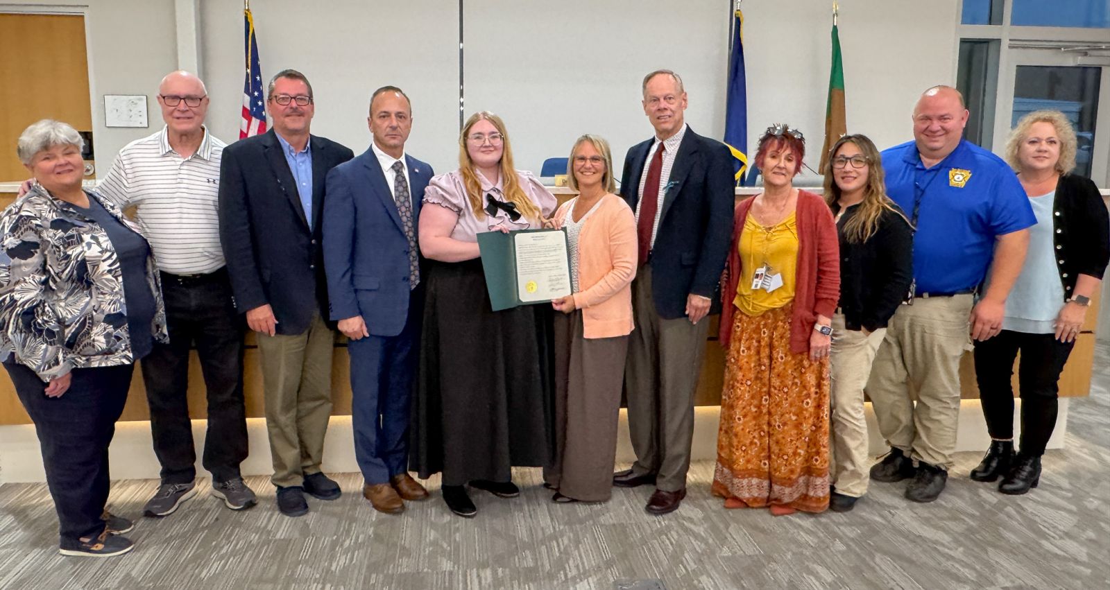 A group of eleven people standing indoors, with a woman in the center holding a framed certificate. Flags are in the background.