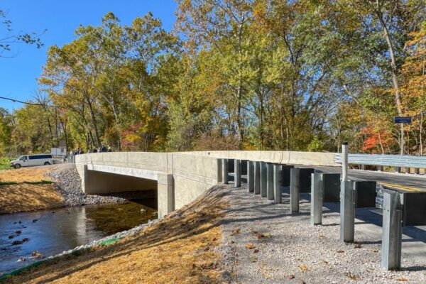 A newly constructed concrete bridge crosses a small creek, surrounded by trees with autumn foliage. A car is parked on the road leading to the bridge under a clear blue sky.